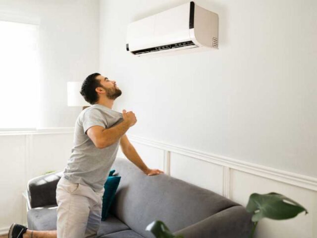 Man holding shirt front open under a wall-mount air conditioning unit in his home.
