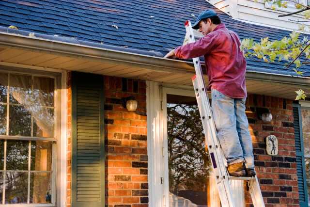 Man cleaning gutters on a ladder against a brick house.