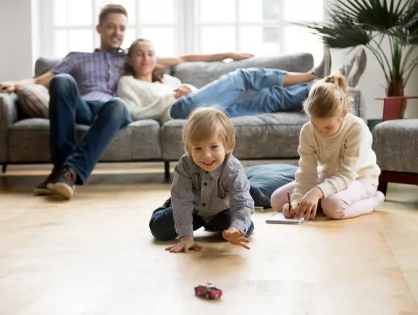 Two children playing on the floor of a living room, watched by parents sitting on a couch.